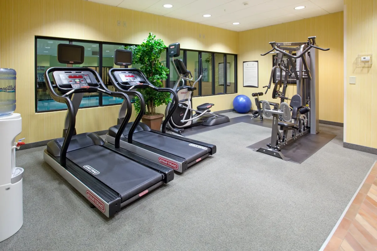 A bright, modern fitness center with treadmills, an elliptical machine, a weight training station, a water cooler, and a fitness ball. The space is designed for solo workouts or small group fitness classes, with a motivational atmosphere.