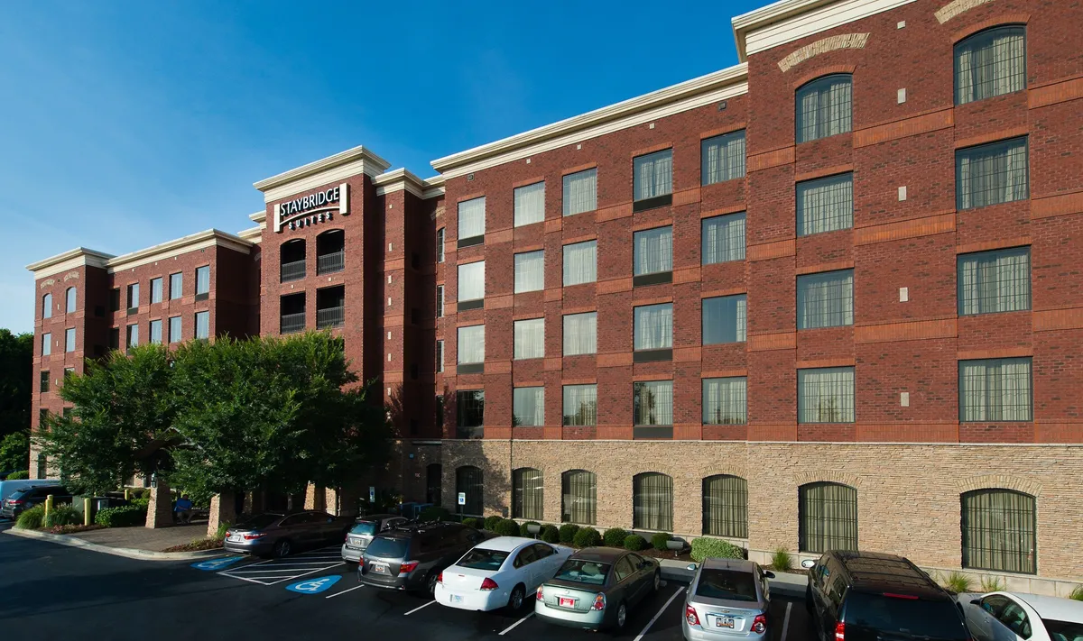 The exterior shot showcases the property's large brick facade, a prominent Staybridge Suites sign, and a well-maintained parking area in the foreground. The scene exudes comfort and accessibility, reinforcing the idea of stress-free arrivals and departures.