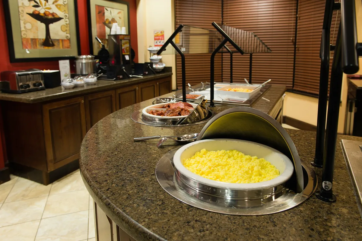 A bright breakfast area featuring a buffet counter lined with yellow scrambled eggs, a well-equipped coffee station, and decorative artwork. The setup suggests a space where guests can fuel up before a busy campus day or unwind with a cup of coffee.