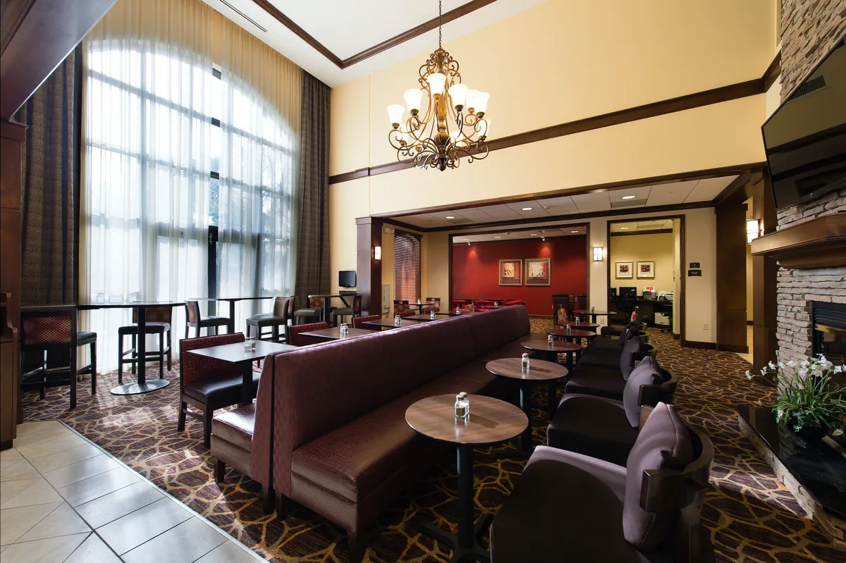 A comfortable living area is depicted with large windows, elegant drapes, a stylish chandelier, a cozy fireplace, and plush couches. The arrangement creates a warm and inviting atmosphere ideal for both relaxation and productivity, with natural light illuminating the space.