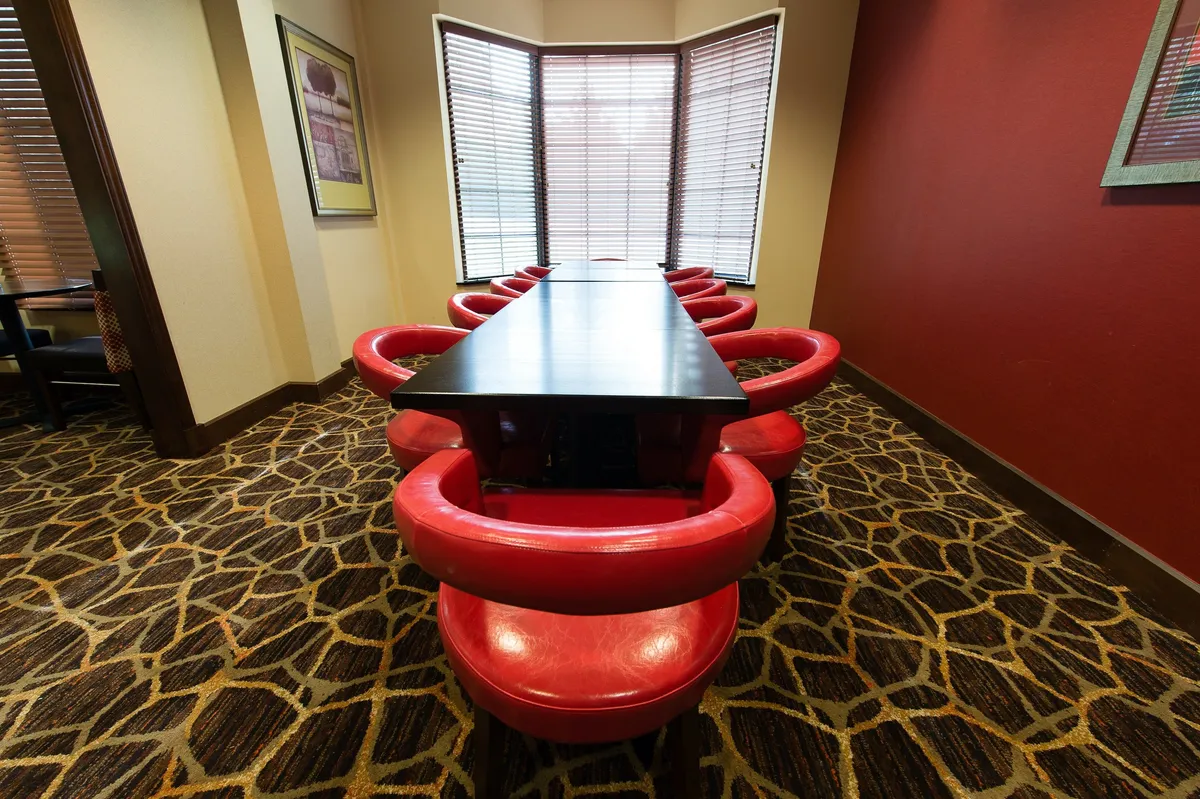 A modern interior featuring a long black table surrounded by vibrant red chairs, situated next to a large window that allows natural light to flood the space. The setup is ideal for business meetings, social gatherings, or brainstorming sessions, and the spacious arrangement invites collaboration and conversation.
