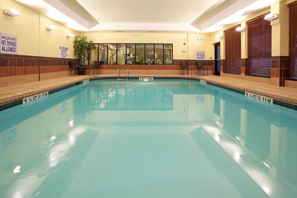 A spacious interior scene featuring a sparkling swimming pool, adjacent fitness area, and large windows that flood the space with natural light. People are seen swimming, lounging poolside, and exercising, creating a lively atmosphere focused on relaxation, health, and connection.