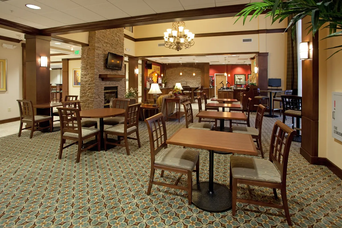 A cozy common area with wooden tables and chairs, a glowing fireplace, a chandelier overhead, vibrant green plants, and decorative wall art. The layout invites guests to gather for casual dining, socializing, or relaxing while watching TV—activities that suit both group celebrations and quiet moments.