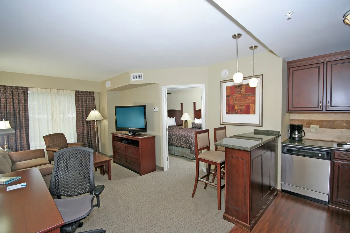 The image showcases a spacious, inviting condo interior with a comfortable sofa facing a large TV, a modern kitchen area, a dining table with chairs, and an open view into the bedroom. The space feels bright, homey, and perfect for unwinding or spending quality time with loved ones.