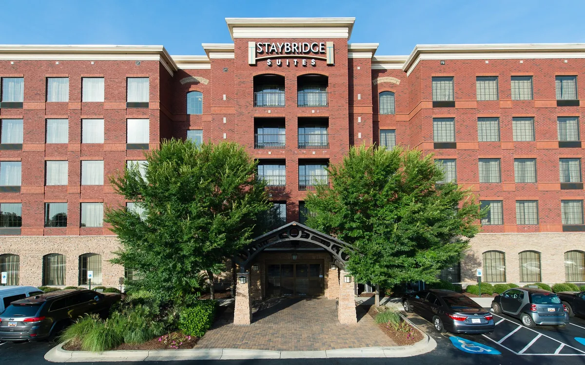 Exterior view of the Staybridge Suites Columbia entrance, featuring a welcoming awning, large windows revealing a glimpse into the lobby, a classic brick facade, and well-maintained landscaping. The scene conveys an inviting point of arrival for guests.