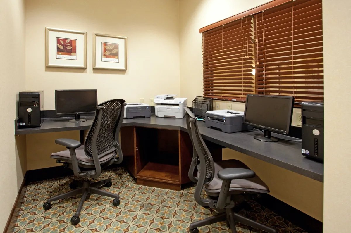 Bright and modern business center with multiple desks, computers, printers, tasteful artwork, and window blinds. The setting is professional, organized, and ready for guests to work productively or prepare for meetings and conferences.