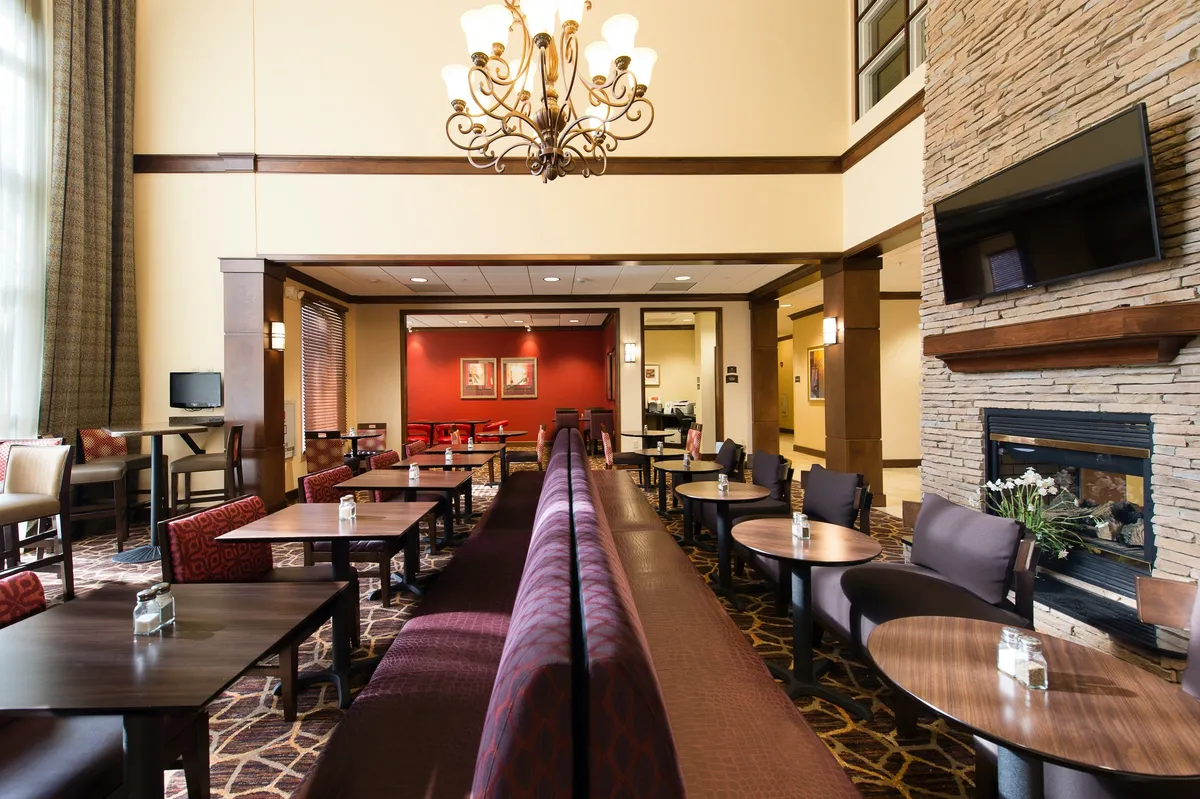 A cozy lounge area featuring a chandelier, inviting fireplace, comfortable seating arrangements, and wall art. Guests are seen socializing, reading, and enjoying refreshments, evoking a warm, communal atmosphere ideal for evening receptions.